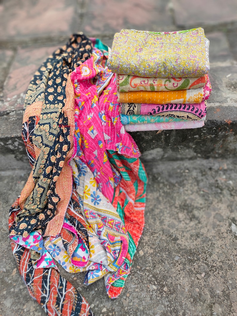 May include: A stack of colorful, patterned quilts and a folded quilt. The quilts feature various designs, including floral and geometric patterns, in shades of pink, yellow, orange, and black. The quilts are made of soft fabric.