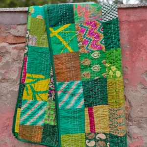 May include: A patchwork quilt made from various fabrics in shades of green, yellow, and brown. The quilt is stitched together with white thread and has a fringed edge.