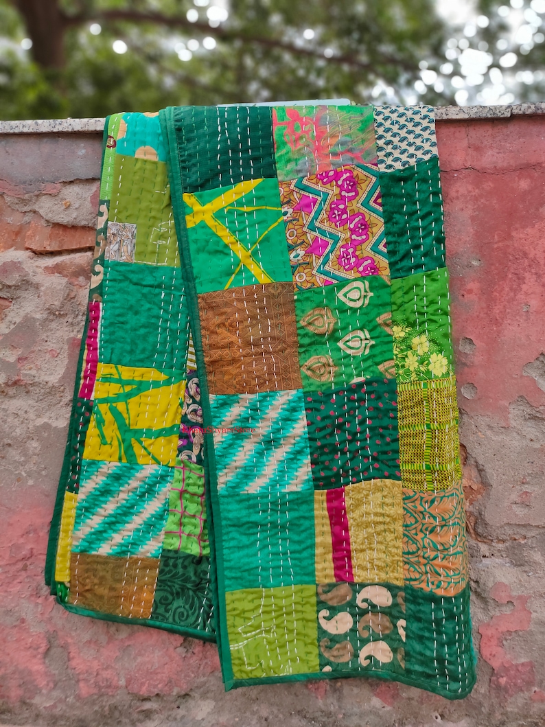May include: A patchwork quilt made from various fabrics in shades of green, yellow, and brown. The quilt is stitched together with white thread and has a fringed edge.
