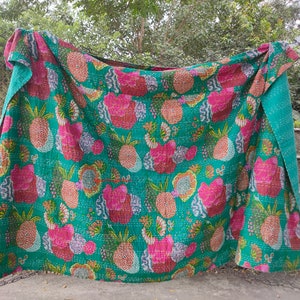 Op de afbeelding: Een turquoise kantha-deken met een roze en oranje ananasprint. De deken is gevouwen en hangt aan een hek.