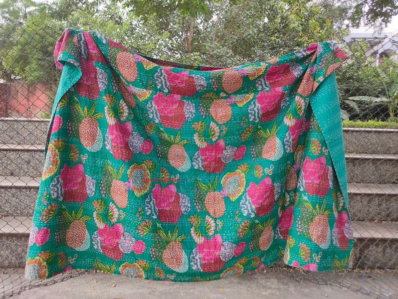 Op de afbeelding: Een turquoise kantha-deken met een roze en oranje ananasprint. De deken is gevouwen en hangt aan een hek.