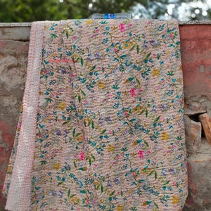 May include: A beige kantha quilt with a floral pattern in shades of pink, blue, green, and yellow. The quilt is folded and draped over a wall.