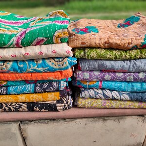May include: Two stacks of colorful, patterned fabric items, possibly quilts or throws. The left stack features green, blue, orange, and black designs. The right stack has tan, green, gray, purple, and blue patterns. The items are stacked on a stone surface.