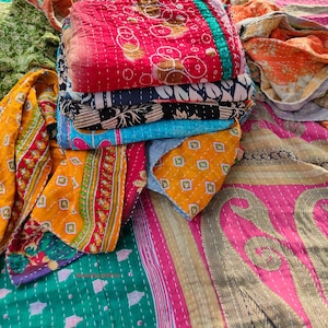 May include: A pile of vibrant kantha quilts with diverse patterns. The quilts display floral, geometric, and abstract designs in red, blue, green, orange, and yellow hues. The quilts are folded and stacked, showcasing their intricate stitching and varied textures.