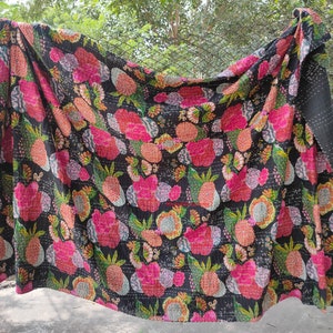 Op de afbeelding: Een zwarte kantha-deken met een bloemen- en ananaspatroon in tinten roze, groen en geel. De deken is gemaakt van katoen en met de hand gestikt.