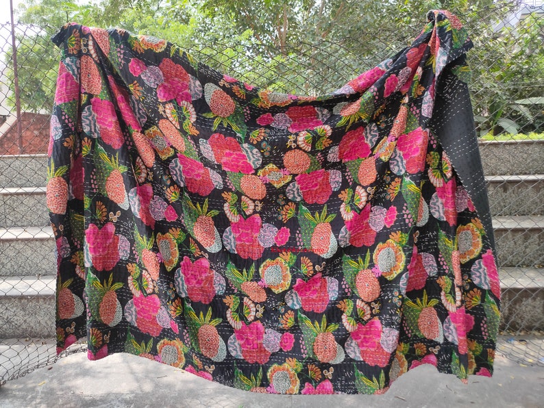 Op de afbeelding: Een zwarte kantha-deken met een bloemen- en ananaspatroon in tinten roze, groen en geel. De deken is gemaakt van katoen en met de hand gestikt.