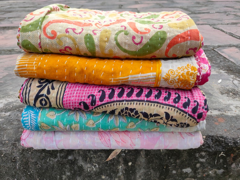 May include: A stack of five colorful, patterned quilts. The top quilt has a beige background with orange, green, and pink paisley designs. The quilts below are yellow, pink, teal, and light pink, each with unique patterns and stitched details.