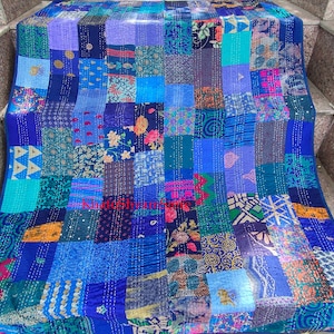 Può includere: Un quilt patchwork realizzato con diversi tessuti blu, viola e verdi. Il quilt è composto da molti quadrati di tessuto diversi, ognuno con un motivo unico. Il quilt è piegato e disposto su una serie di gradini in pietra.