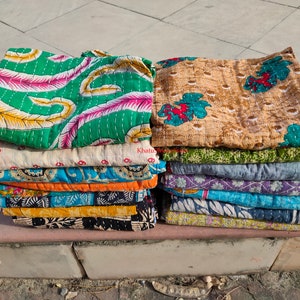 May include: Stacks of colorful, patterned quilts. The quilts feature various designs, including floral and abstract patterns in shades of green, pink, blue, and yellow. The quilts are folded and stacked on top of each other.