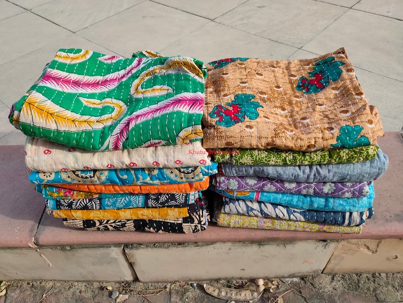 May include: Stacks of colorful, patterned quilts. The quilts feature various designs, including floral and abstract patterns in shades of green, pink, blue, and yellow. The quilts are folded and stacked on top of each other.