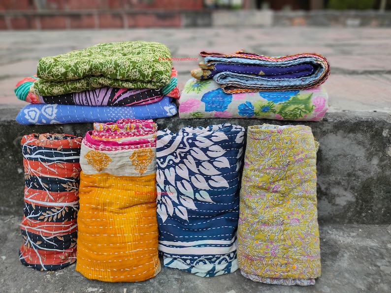 May include: A collection of colorful, patterned kantha quilts. The quilts are stacked and rolled, showcasing various designs including floral, leaf, and geometric patterns in shades of blue, green, yellow, orange, and pink. The image conveys a handcrafted, textile theme.