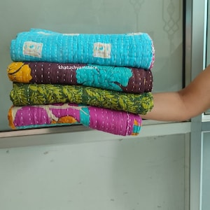 Könnte beinhalten: Ein Stapel von vier Kantha-Steppdecken in verschiedenen Farben und Mustern. Die oberste Steppdecke ist türkis mit einem weißen geometrischen Muster. Die zweite Steppdecke ist braun mit einem türkisfarbenen Blumenmuster. Die dritte Steppdecke ist grün mit einem rosa Blumenmuster. Die unterste Steppdecke ist rosa mit einem gelben Blumenmuster. Die Steppdecken sind gefaltet und übereinander gestapelt. Der Text "khatushyamstore" ist auf der zweiten Steppdecke sichtbar.