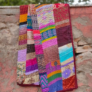 May include: A colorful patchwork quilt made from repurposed fabric scraps. The quilt is made up of squares of different colors and patterns, including floral, paisley, and geometric designs. The quilt is edged in a dark red fabric.