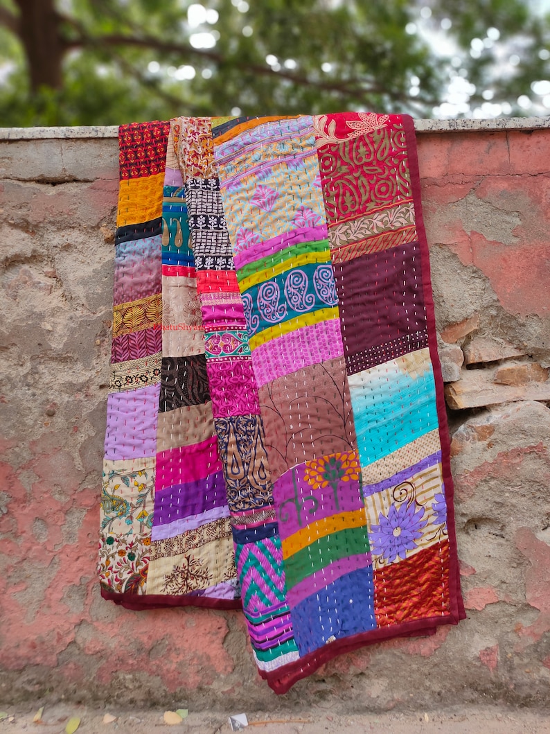 May include: A colorful patchwork quilt made from repurposed fabric scraps. The quilt is made up of squares of different colors and patterns, including floral, paisley, and geometric designs. The quilt is edged in a dark red fabric.