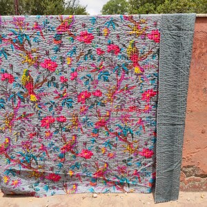 May include: A gray kantha quilt with a floral and bird pattern. The quilt has pink, yellow, blue, and green flowers and birds. The quilt is hanging on a wall.