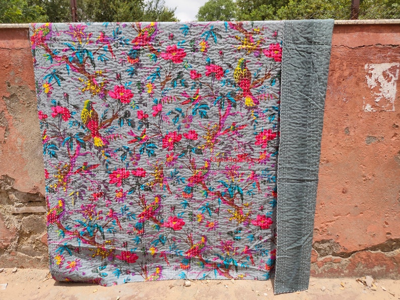 May include: A gray kantha quilt with a floral and bird pattern. The quilt has pink, yellow, blue, and green flowers and birds. The quilt is hanging on a wall.