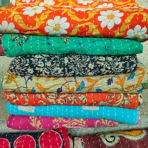 May include: A stack of eight colourful kantha quilts with various floral and geometric patterns. The quilts are made of recycled saris and are hand-stitched. The quilts are folded and stacked on top of each other. The top quilt is orange with white flowers. The bottom quilt is maroon with a paisley pattern. The quilts are all different sizes and shapes. #kanthaquilts #kantha #handmade #recycled #saris #quilts #indianquilts #homedecor #bedding #throwblanket #boho #vintage #ethnic #colourful #patterned #geometric #floral #khatushyamstore