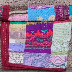May include: A patchwork quilt made from colorful silk fabric scraps. The quilt is hand-stitched and features a variety of patterns and colors, including purple, pink, blue, green, and red. The quilt is folded in half and has a maroon border.