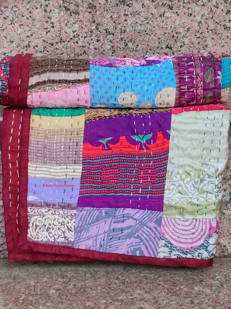 May include: A patchwork quilt made from colorful silk fabric scraps. The quilt is hand-stitched and features a variety of patterns and colors, including purple, pink, blue, green, and red. The quilt is folded in half and has a maroon border.