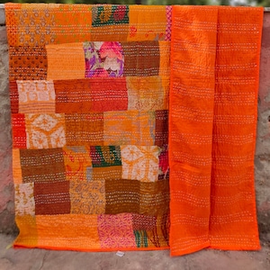 Op de afbeelding: Een patchworkdeken met verschillende oranje, bruine en roze stoffen. De deken is gemaakt van vierkanten en rechthoeken in verschillende maten en patronen. De deken is afgewerkt met een effen oranje stof.