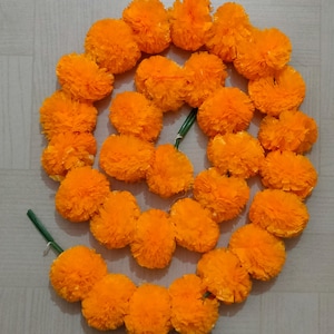 Peut inclure: Une guirlande circulaire de fleurs de souci orange vif. Les fleurs sont densément tassées, créant un aspect plein et texturé. Des tiges vertes sont visibles, reliant les fleurs. La guirlande est disposée sur une surface de couleur claire.