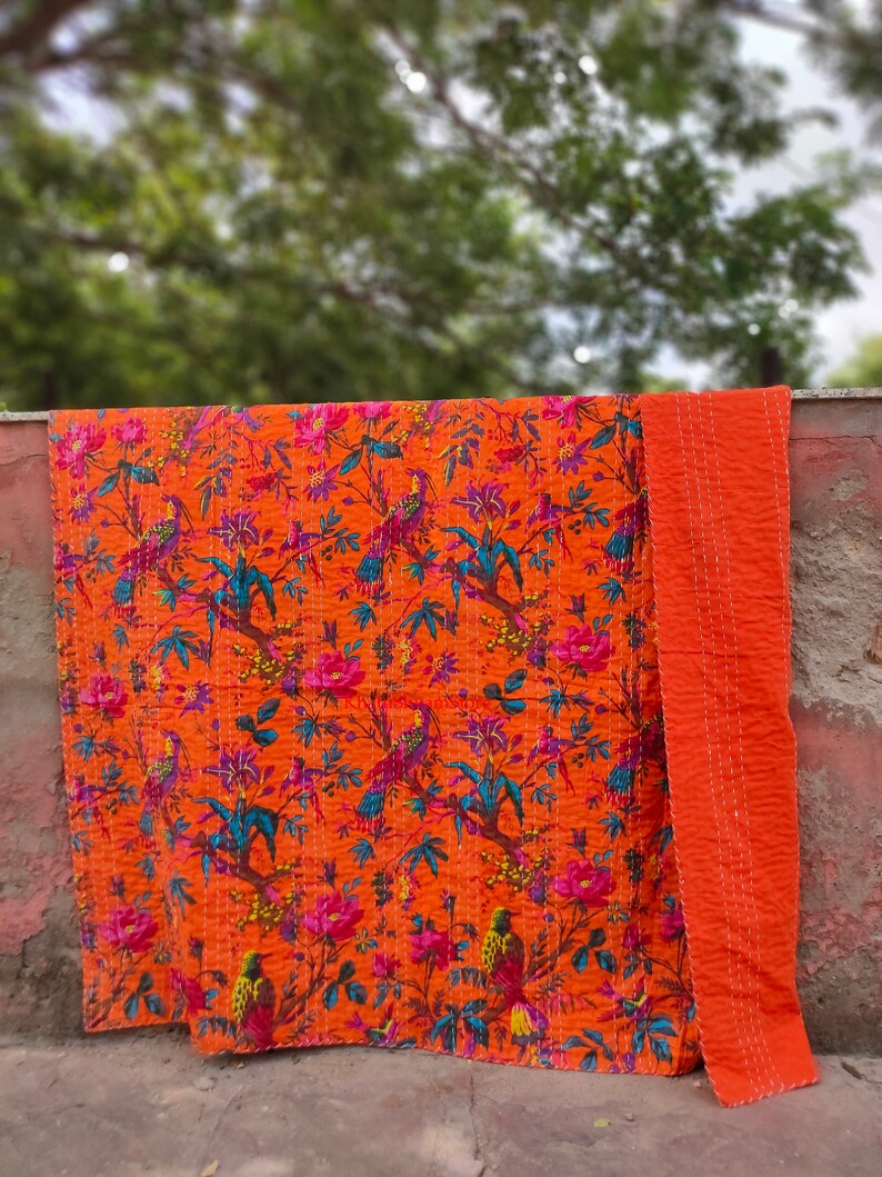 May include: An orange kantha quilt with a floral and bird pattern. The quilt is folded in half and has a solid orange back.