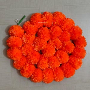 Könnte beinhalten: Eine kreisförmige dekorative Girlande aus zahlreichen leuchtend orangefarbenen Ringelblumen. Die dichten, gerüschten Blütenblätter bilden eine volle, runde Form mit einem kleinen grünen Stiel am oberen linken Rand, was für ein strukturiertes Blumenarrangement sorgt.