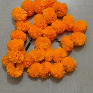 Könnte beinhalten: Eine Girlande aus leuchtend orangefarbenen Ringelblumen. Die Blüten sind dicht gepackt und erzeugen ein volles und strukturiertes Aussehen. Die Girlande ist mit einem grünen Stiel zusammengebunden, ideal für festliche Dekorationen.