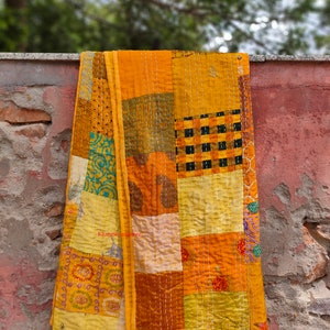 May include: A yellow kantha quilt with a patchwork design. The quilt is made of different fabrics and is hand-stitched with a running stitch. The quilt is hanging on a brick wall.