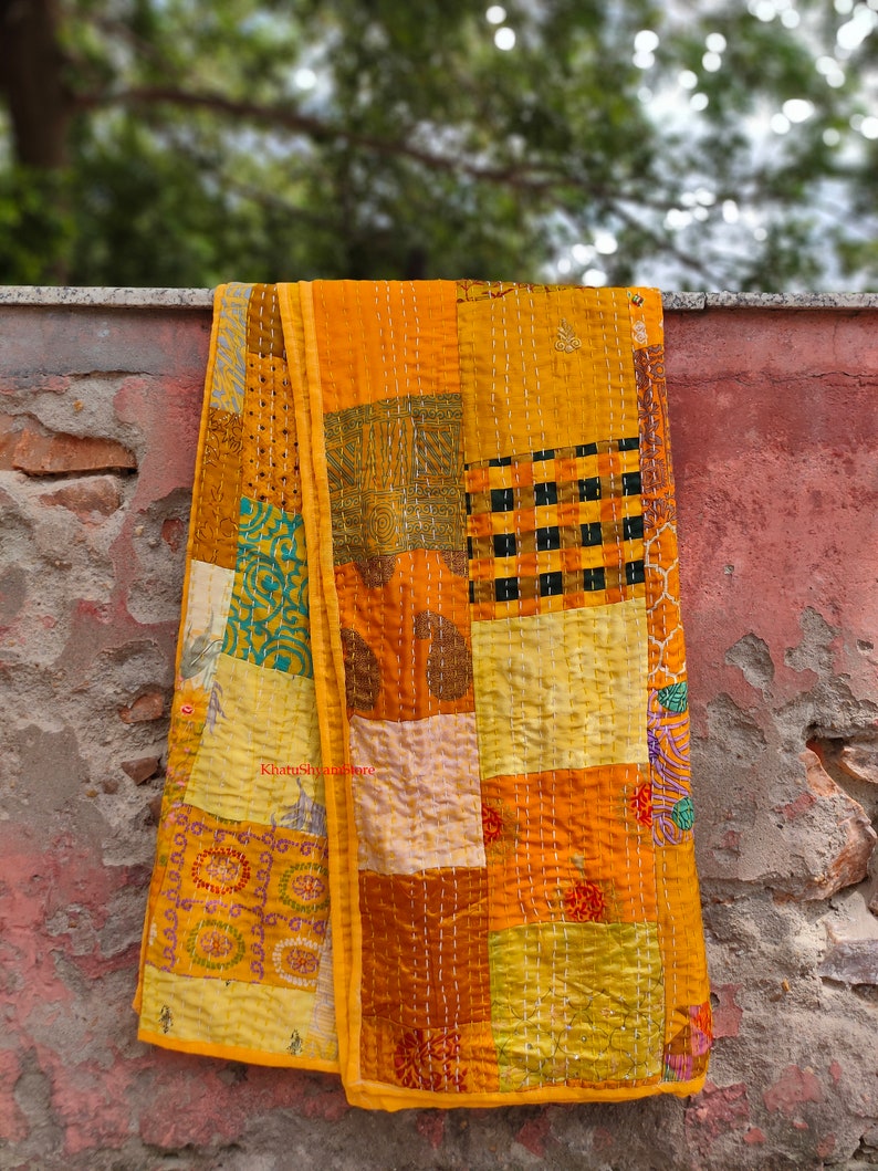 May include: A yellow kantha quilt with a patchwork design. The quilt is made of different fabrics and is hand-stitched with a running stitch. The quilt is hanging on a brick wall.