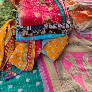 May include: A stack of colorful, quilted textiles in various patterns and colors, including red, blue, yellow, and green. The textiles appear to be handmade with visible stitching and feature floral and geometric designs. The fabrics are folded and stacked together.