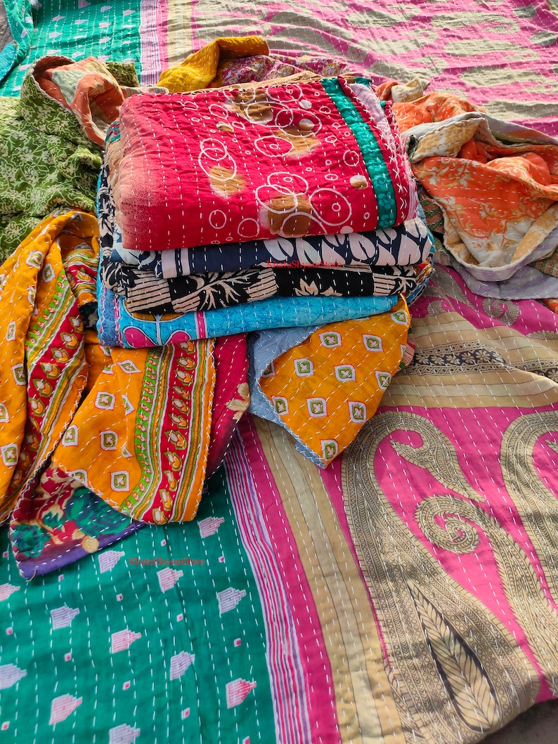 May include: A stack of colorful, quilted textiles in various patterns and colors, including red, blue, yellow, and green. The textiles appear to be handmade with visible stitching and feature floral and geometric designs. The fabrics are folded and stacked together.