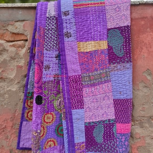 May include: A purple patchwork quilt with a variety of floral and geometric patterns. The quilt is made of different shades of purple, pink, and blue fabrics. It is folded and hanging on a brick wall.