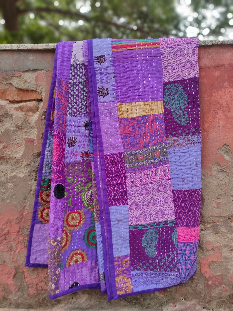 May include: A purple patchwork quilt with a variety of floral and geometric patterns. The quilt is made of different shades of purple, pink, and blue fabrics. It is folded and hanging on a brick wall.