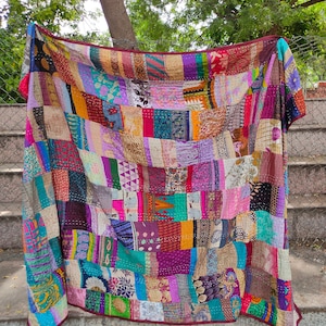 Könnte beinhalten: Eine bunte Patchwork-Decke aus wiederverwendeten Stoffresten. Die Decke ist ein Mix aus leuchtenden Farben und Mustern, darunter florale, geometrische und Paisley-Designs. Die Decke hängt an einem Zaun und hat einen burgunderfarbenen Rand.