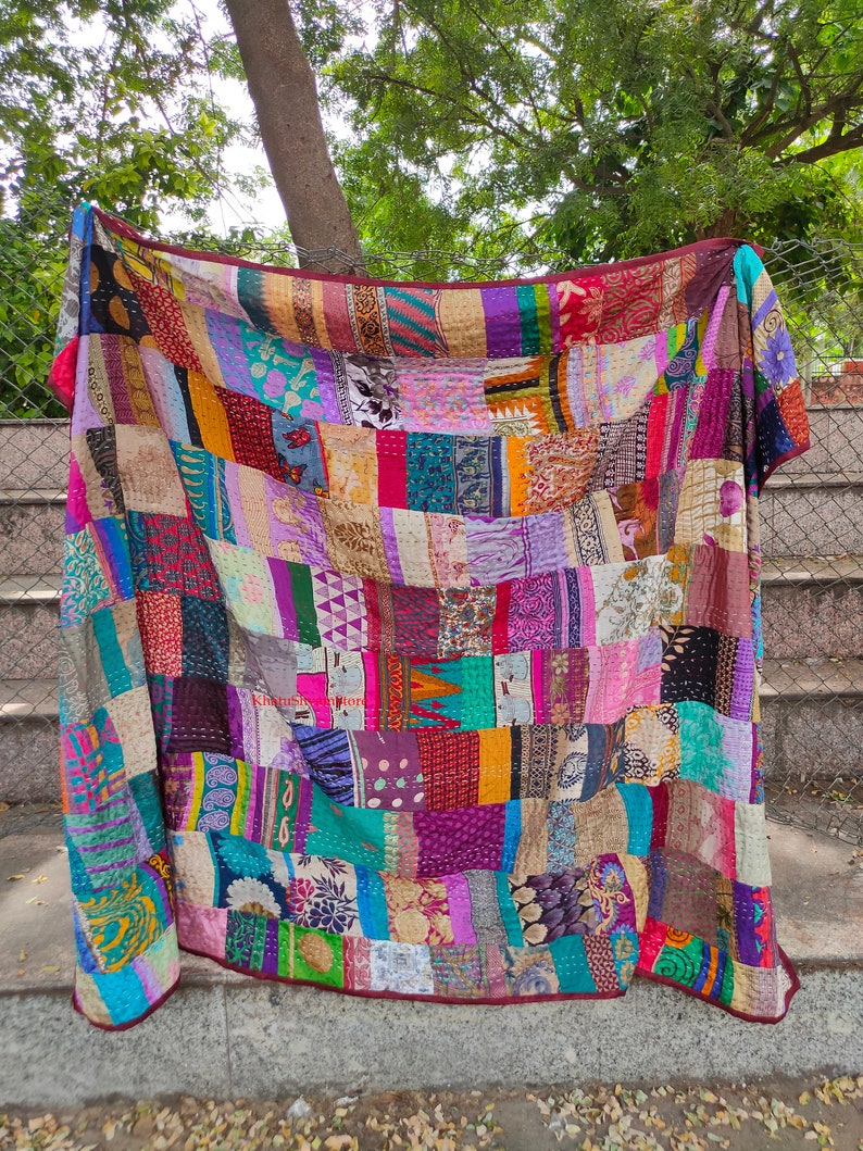 May include: A colorful patchwork quilt made from repurposed fabric scraps. The quilt is a mix of bright colors and patterns, including floral, geometric, and paisley designs. The quilt is hanging on a fence and has a burgundy border.
