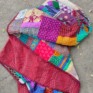 May include: A colorful patchwork quilt made from repurposed fabric scraps. The quilt is made up of squares of different colors and patterns, including purple, red, green, blue, and yellow. The quilt is folded in half and lying on a concrete surface.