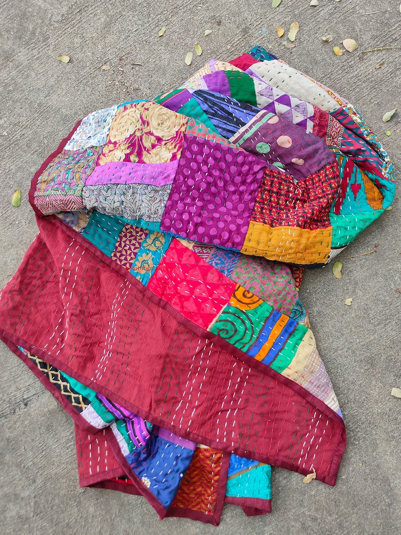 May include: A colorful patchwork quilt made from repurposed fabric scraps. The quilt is made up of squares of different colors and patterns, including purple, red, green, blue, and yellow. The quilt is folded in half and lying on a concrete surface.