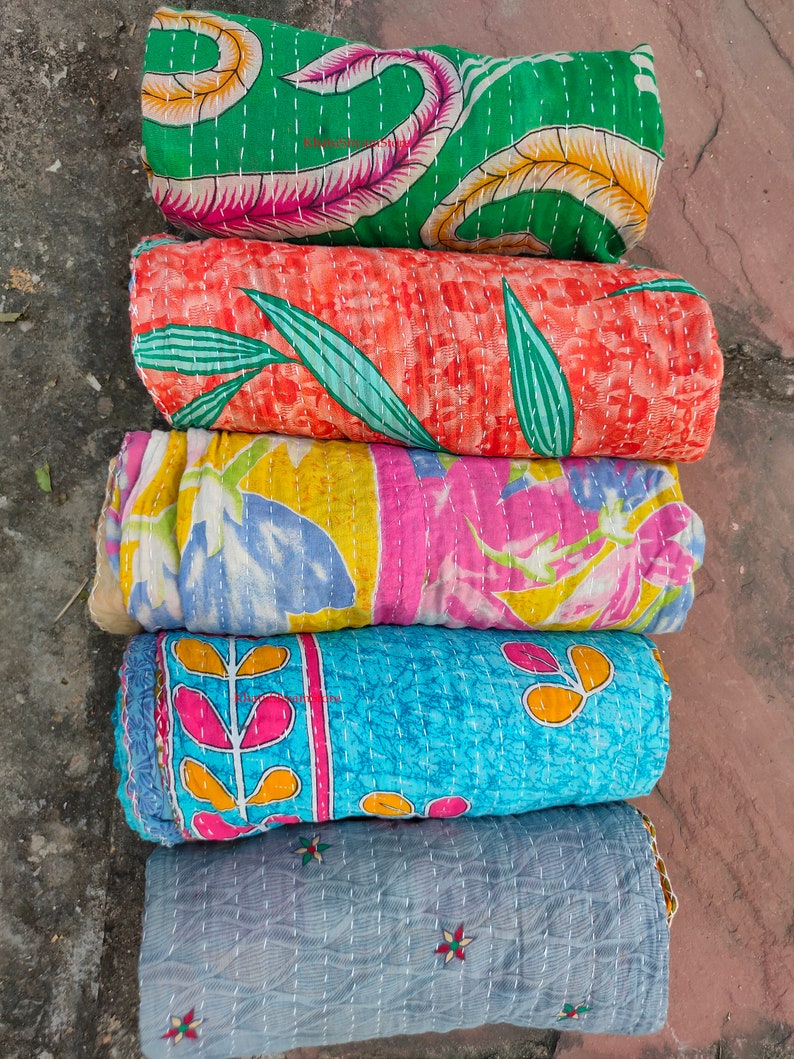 May include: Five rolled, colorful, quilted fabric pieces. The top piece is green with a floral pattern, followed by orange with leaves, then pink and yellow, blue with a floral design, and finally, a gray piece with embroidered flowers.