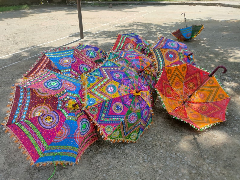 May include: A group of colorful, patterned umbrellas with intricate embroidery and mirror embellishments. The umbrellas are arranged on a gray, textured surface.