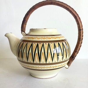 May include: Cream ceramic teapot with a woven handle. The teapot has a geometric pattern in brown, black, and yellow. The handle is made of woven brown material. The spout is on the left side of the teapot.
