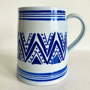 May include: A white ceramic mug with a blue and white geometric pattern. The mug has a handle and a wide rim.