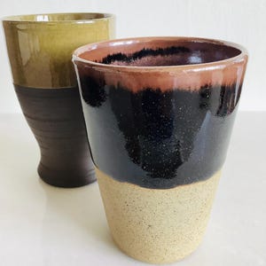 May include: Two handmade ceramic mugs. One is olive green over dark brown, the other is black over tan. The mugs have a rustic, textured appearance, with a glossy finish on the upper portions. The mugs are likely for beverages.
