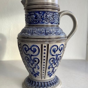 May include: A large, grey stoneware jug with blue and brown accents. The jug has a wide base and a tall, slender body. It is decorated with intricate floral and geometric patterns.