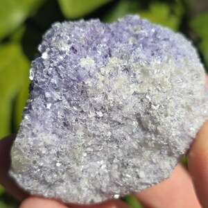 May include: A close-up of a rough, natural fluorite crystal. The stone has a mix of light purple and white colors, with a crystalline texture. The surface reflects light, creating a sparkling effect. The background is blurred green.