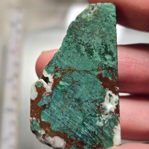 May include: A close-up of a rough-cut mineral specimen. The stone features a vibrant teal-green hue with streaks of brown and patches of white. The texture appears to be slightly rough and uneven, showcasing natural variations.