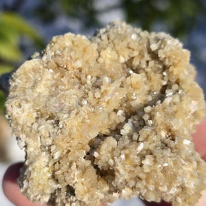 May include: A close-up of a cluster of light yellow crystals with a sparkling, textured surface. The crystals have a layered appearance, with some areas reflecting light. The overall shape is irregular and natural, suggesting a mineral specimen.