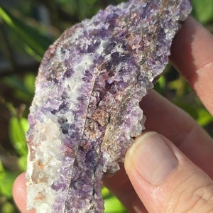 May include: A close-up of a raw, natural amethyst crystal. The stone displays a mix of purple, white, and brown hues, with a rough, textured surface. The crystal is held in a hand, showcasing its natural form.