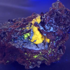 May include: A close-up of a mineral specimen with a rough, textured surface. The rock is primarily reddish-brown with patches of bright yellow and green. The background is a dark blue.