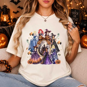 Könnte beinhalten: Ein cremefarbenes T-Shirt mit einem Grafikdruck verschiedener Zeichentrickfiguren in Halloween-Kostümen, die einen zentralen ovalen Rahmen mit einer Spukhaus-Illustration umgeben, akzentuiert durch orangefarbene und schwarze Luftballons und Kürbisse.